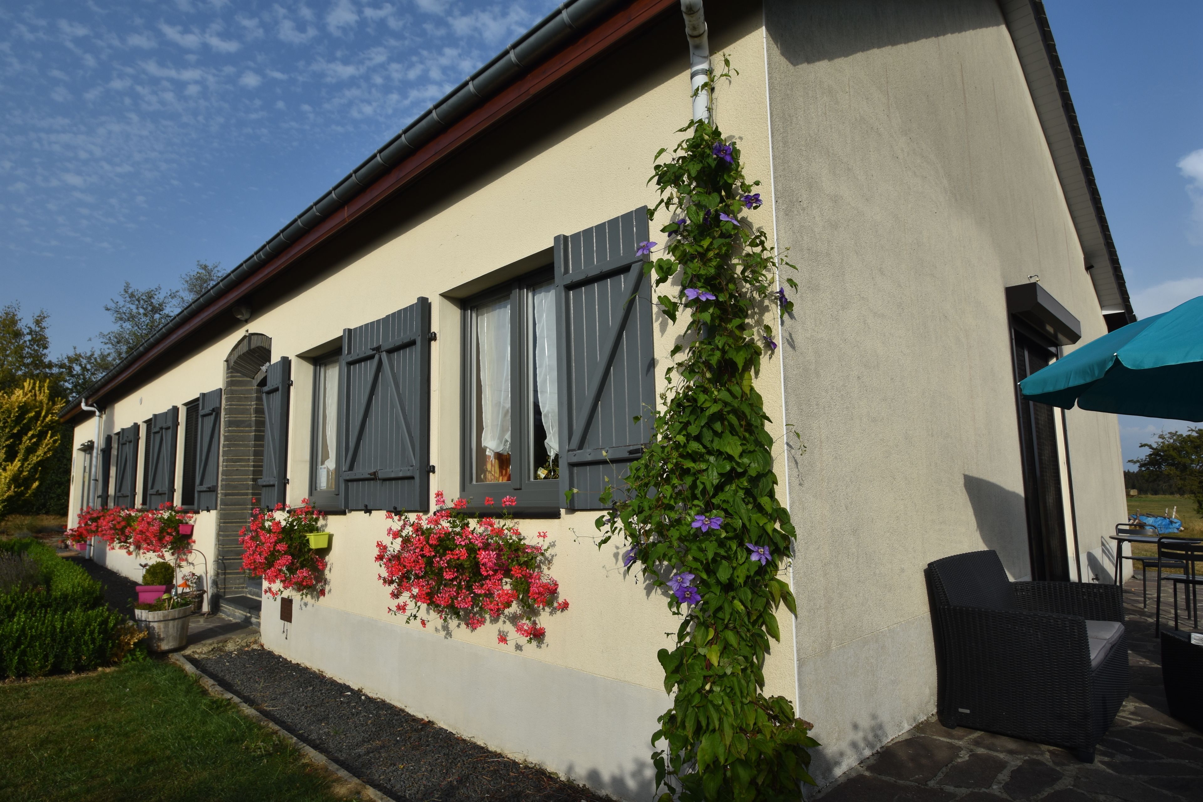 Un edificio de fachada color beige, contraventanas grises y una planta trepadora verde. Hay jardineras con flores rosas y rojas en la planta baja.