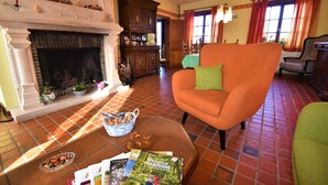 Een woonkamer met een open haard, een grote oranje fauteuil, een salontafel met tijdschriften en een eethoek op de achtergrond.