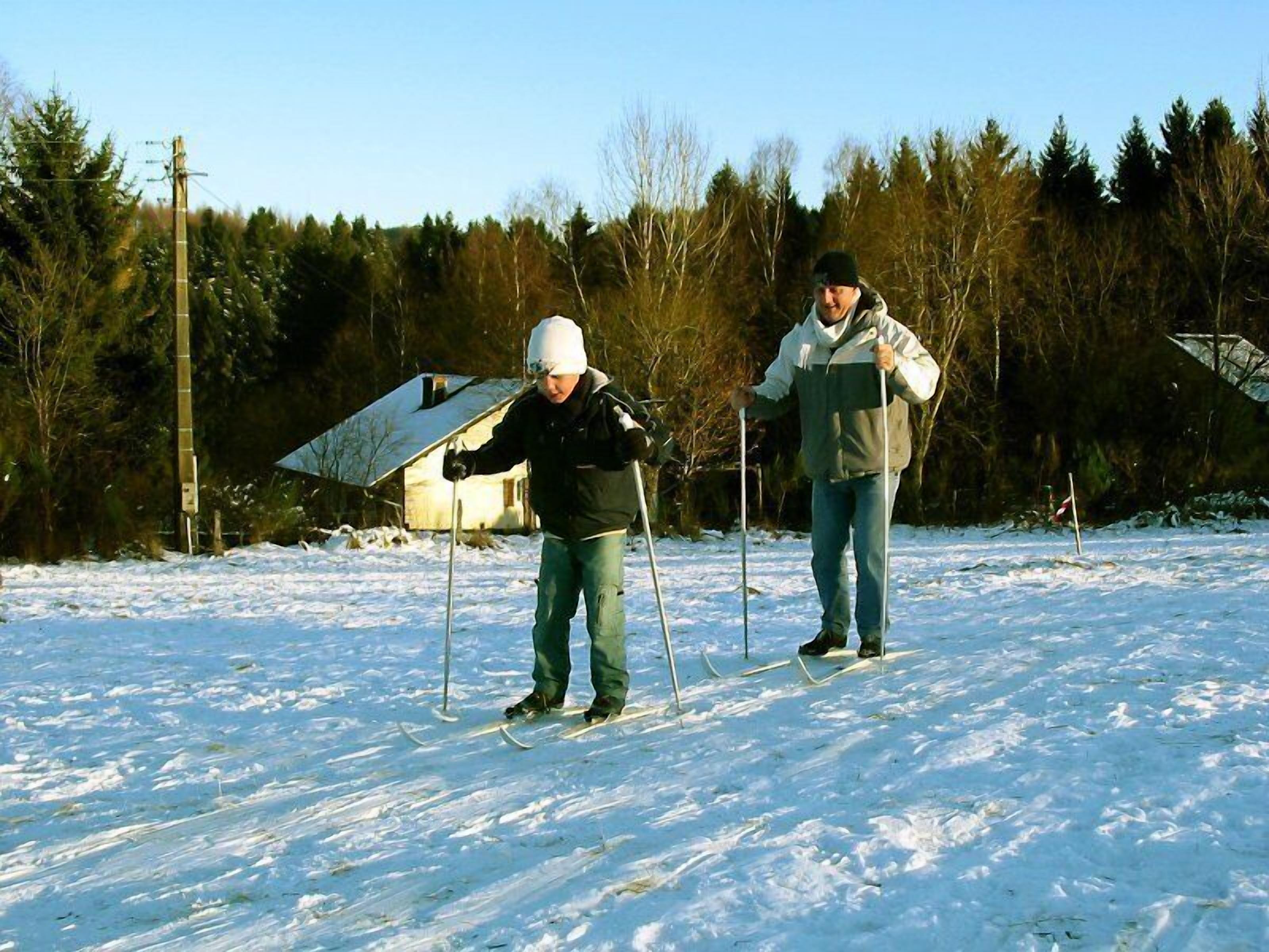Two people cross-country skiing on a snowy field with a house and trees in the background.