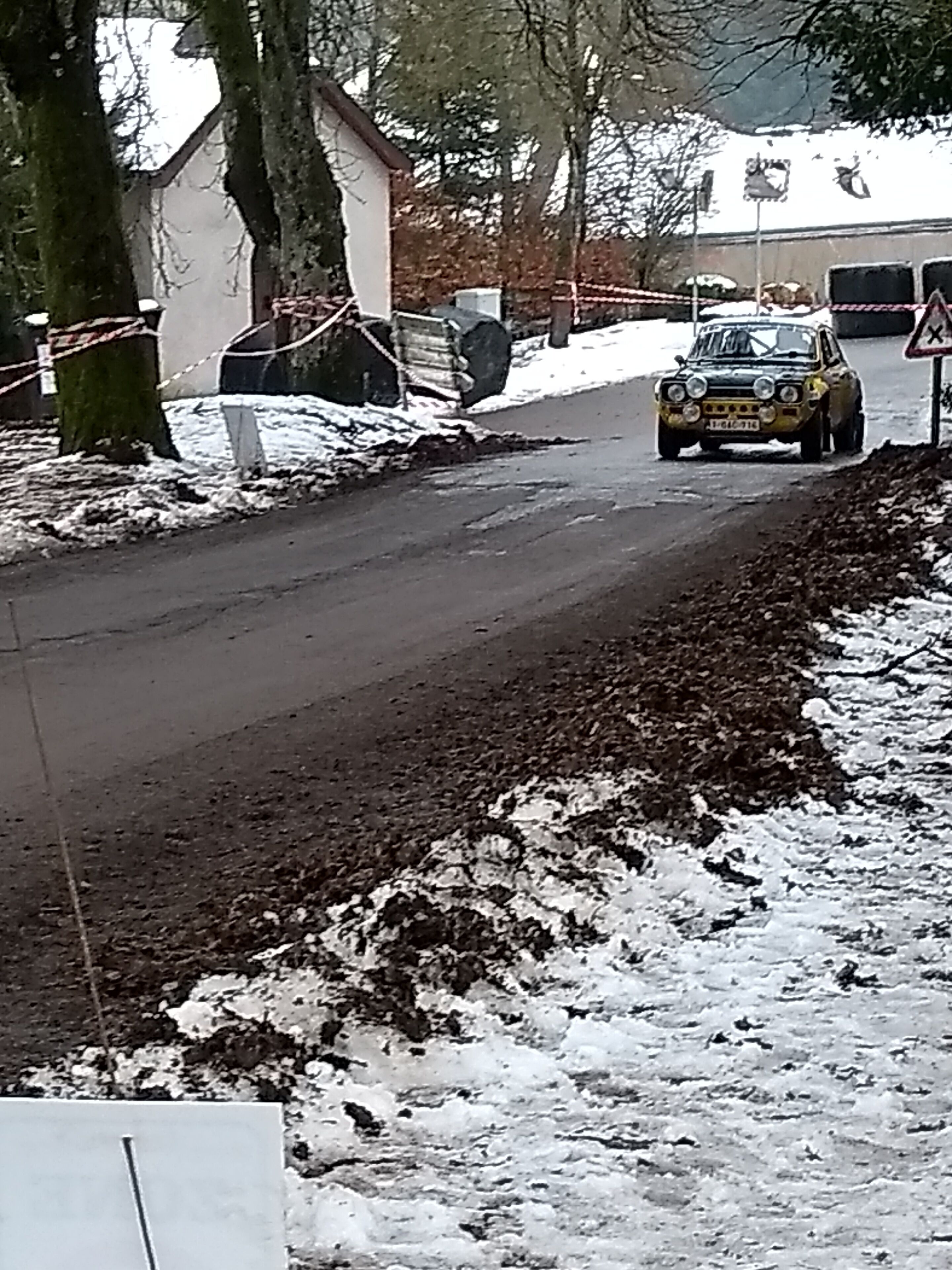 Una carretera rural con un coche de rally amarillo, árboles y un edificio al fondo.