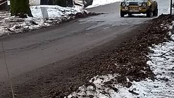Una carretera rural con un coche de rally amarillo, árboles y un edificio al fondo.
