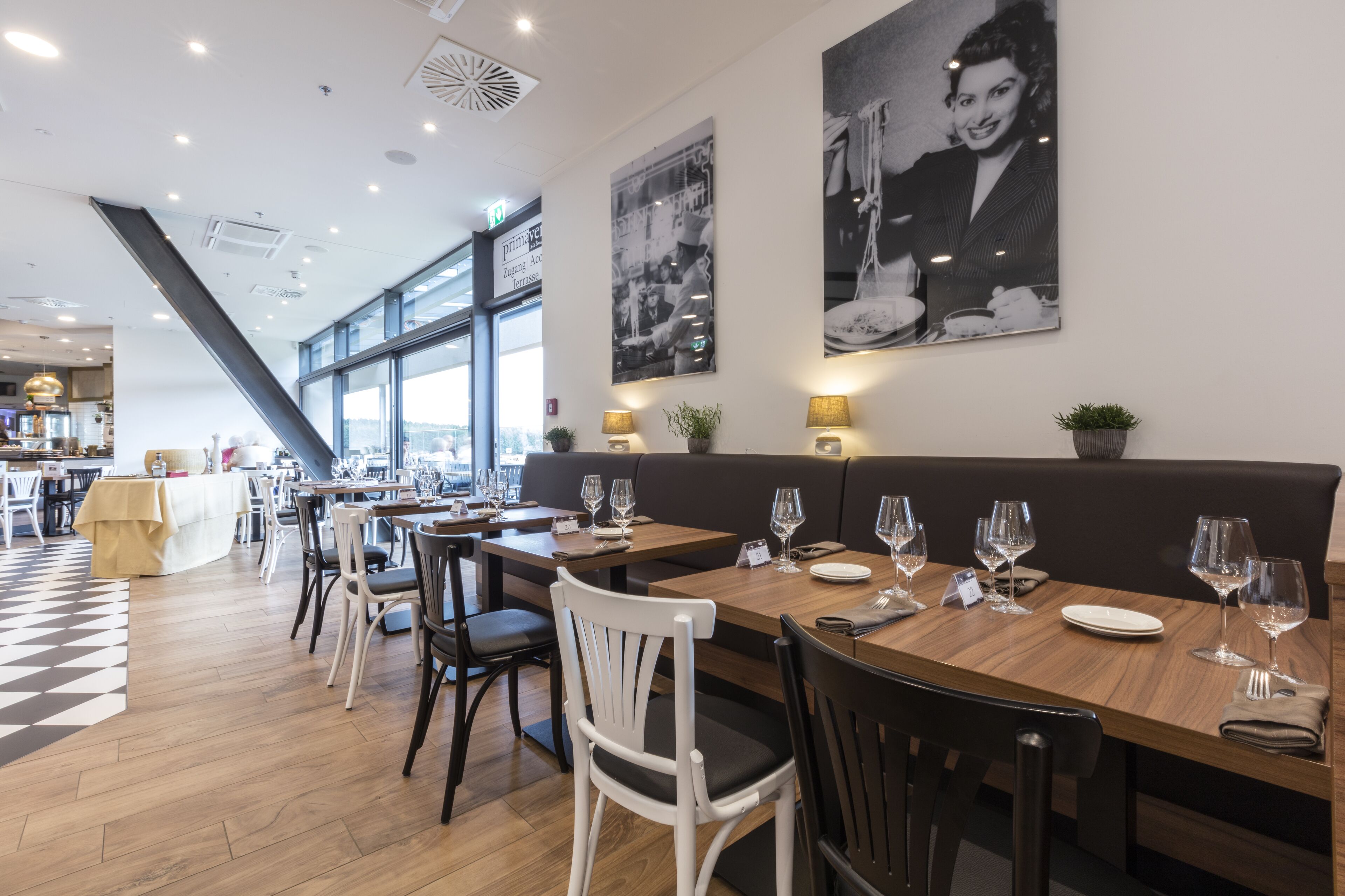 A restaurant with wooden tables and chairs, large windows, and black-and-white photographs on the walls.