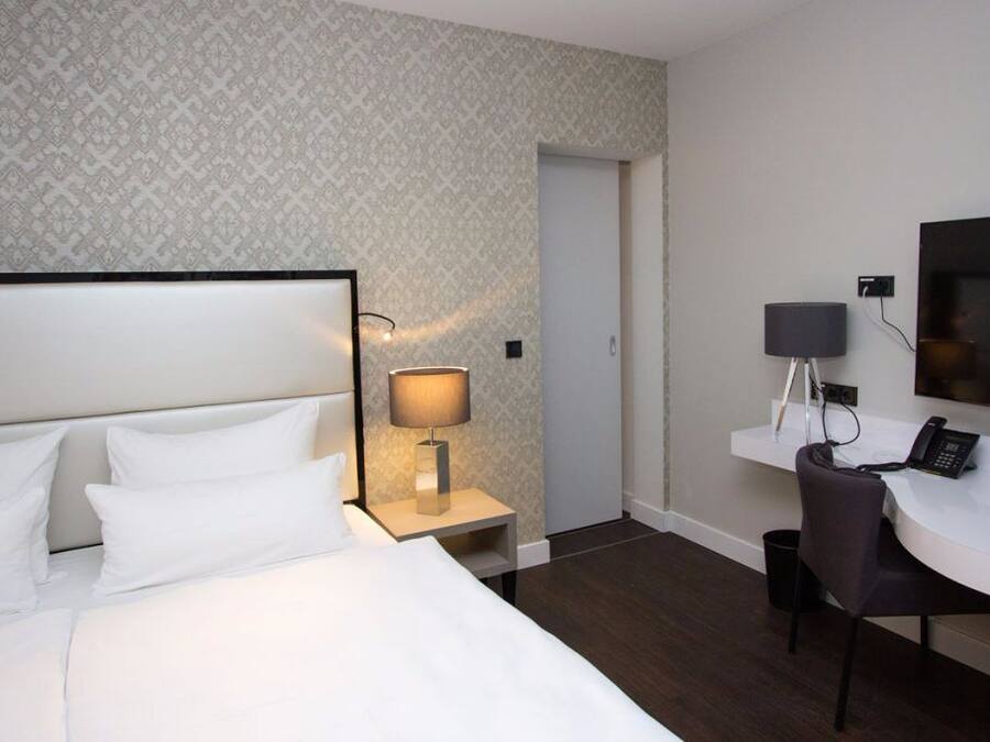 A hotel room with a bed, a desk, a chair, a lamp, a TV, and a patterned wallpaper.
