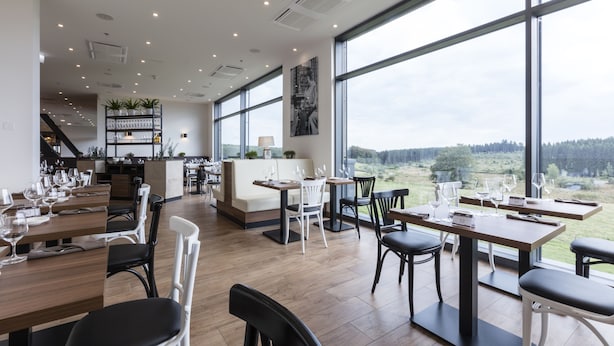 A modern restaurant with large windows offering a view of the outdoors. Tables are set with wine glasses and plates.