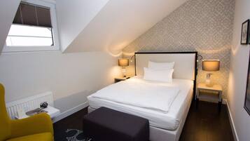 Ein Schlafzimmer mit einem Bett, zwei Nachttischlampen, einem Fenster mit Jalousien, einem Heizkörper, einem gelben Sessel und einem schwarzen Hocker.