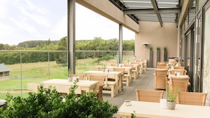 An outdoor patio with wooden tables and wicker chairs, overlooking a grassy area with trees.