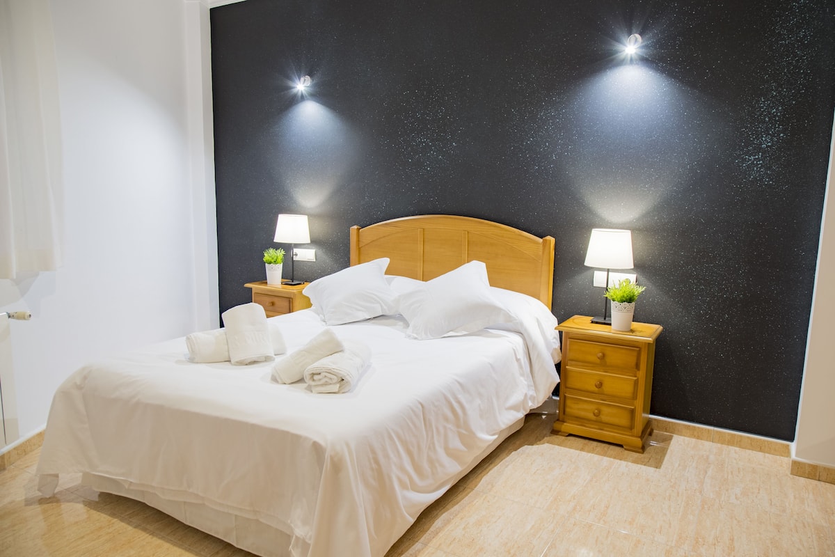 A neatly made bed with white linens and pillows, two bedside tables with lamps and small plants, and a starry night wall.