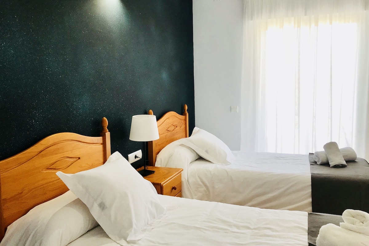 A hotel room with two beds, a nightstand, a lamp, and a window with curtains.