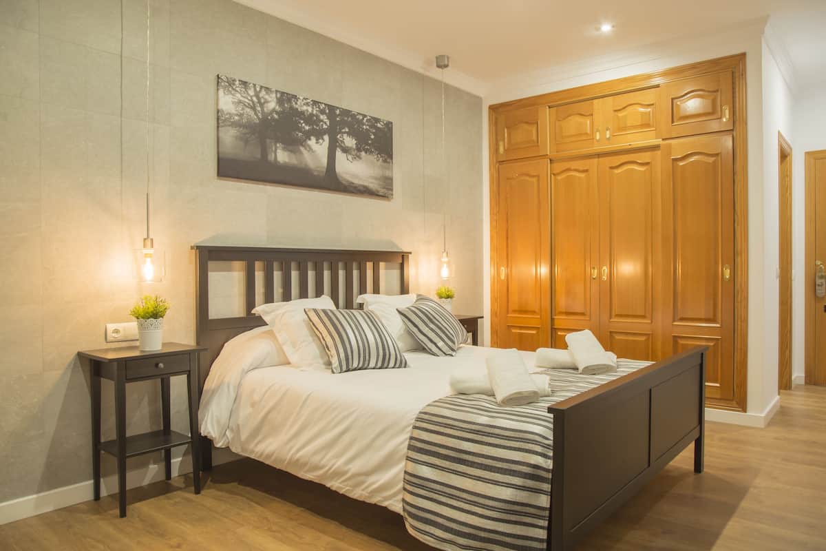 A neatly made bed with a striped bedspread, a bedside table with a plant, and a wall-mounted painting.