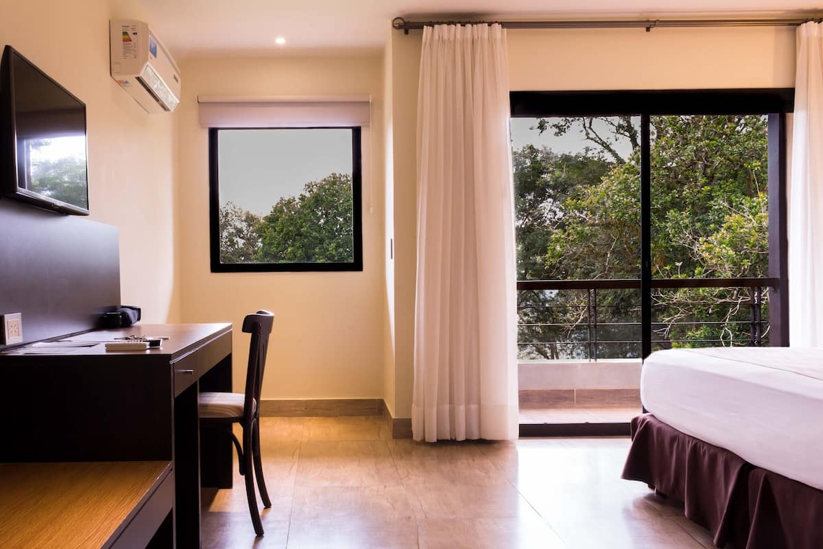 A hotel room with a bed, a desk, a chair, a TV, a window, and an air conditioning unit.