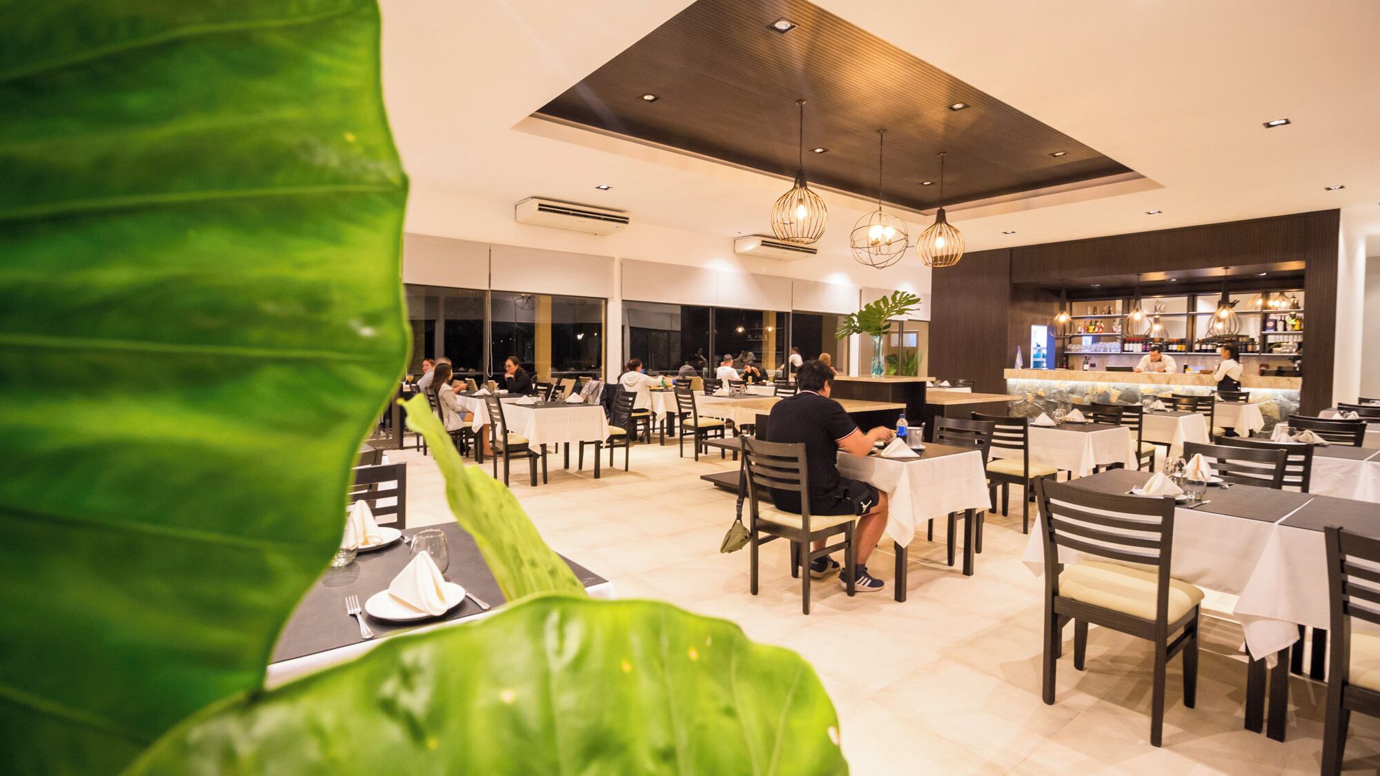 A modern restaurant with multiple tables and chairs, large windows, and hanging pendant lights.