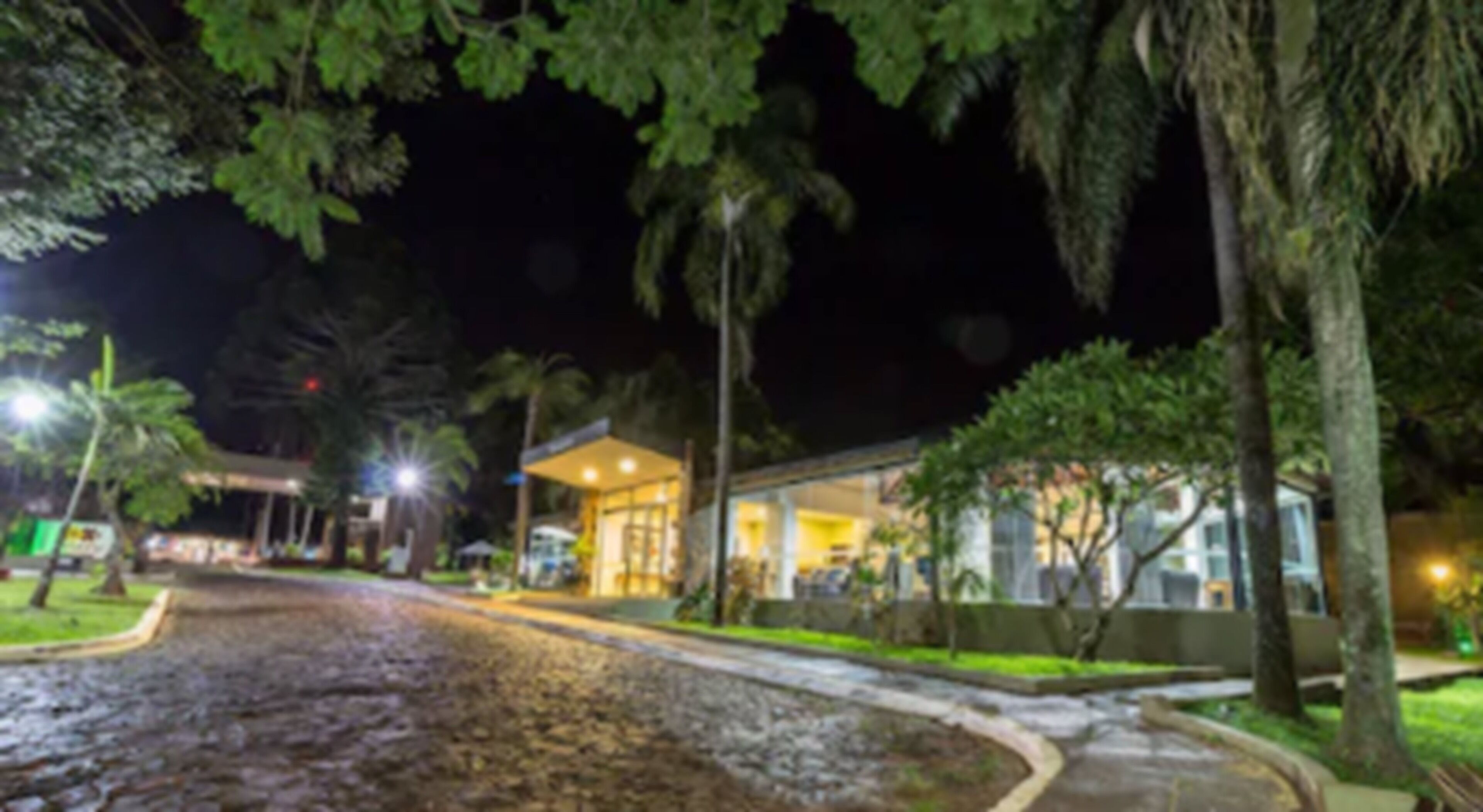 Vista nocturna de un hotel con acceso pavimentado, un espacio de estar techado al aire libre y vegetación circundante.