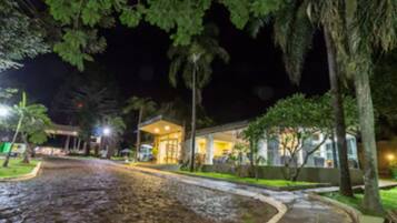 Vista nocturna de un hotel con acceso pavimentado, un espacio de estar techado al aire libre y vegetación circundante.