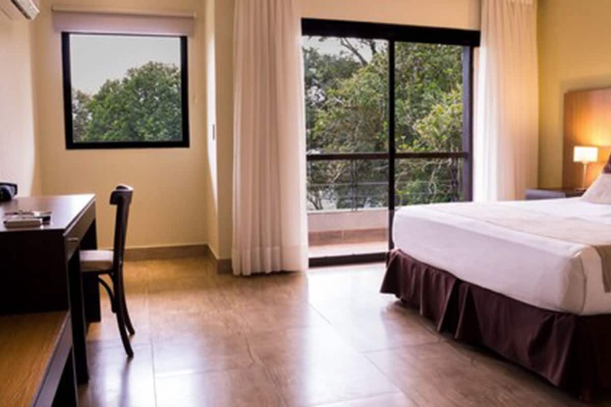 A hotel room with a bed, a desk, and a window with a view of trees.