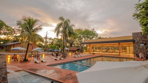 Un área de piscina con sillones reclinables y sombrillas, un restaurante con terraza y un edificio con amplias ventanas.