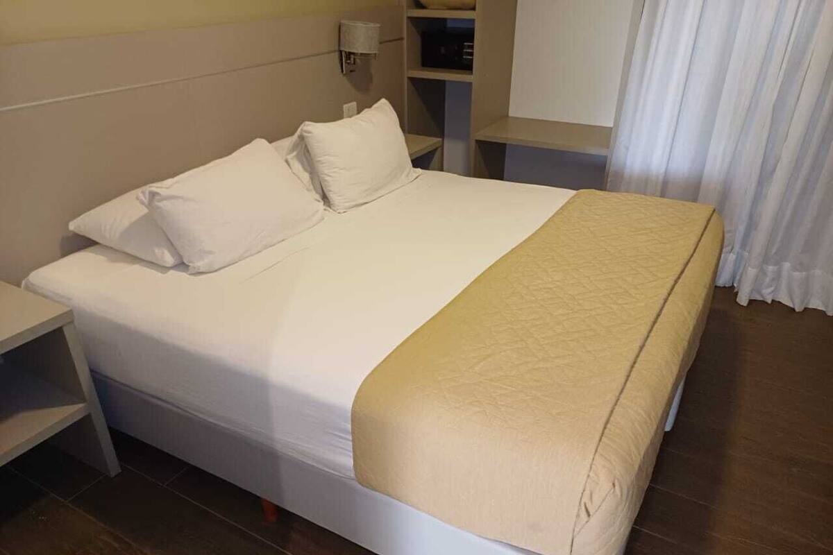 A neatly made bed with white bedding and a beige throw blanket, a bedside table, and a lamp.
