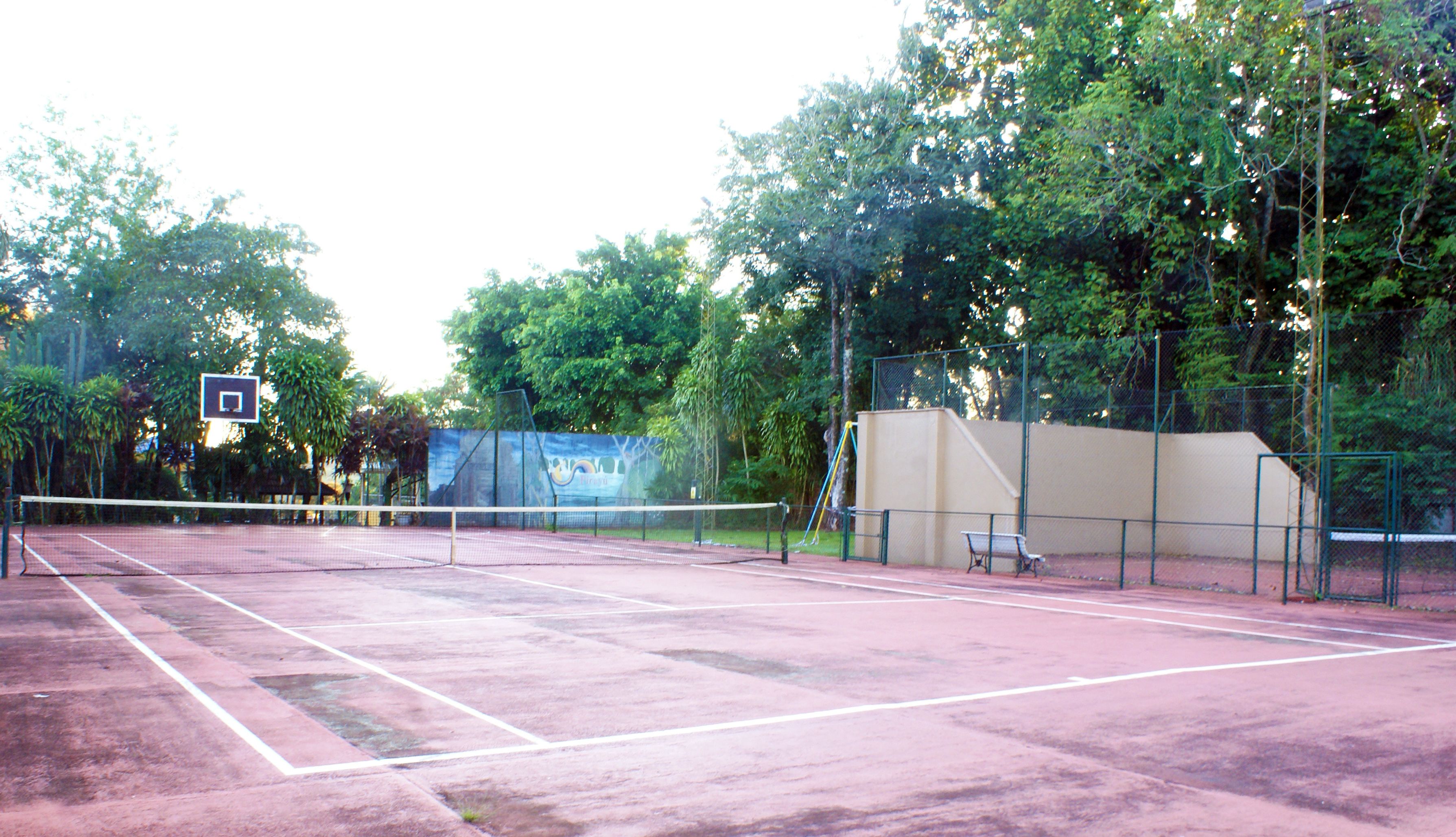 An outdoor tennis court with a net, a basketball hoop, and a bench.