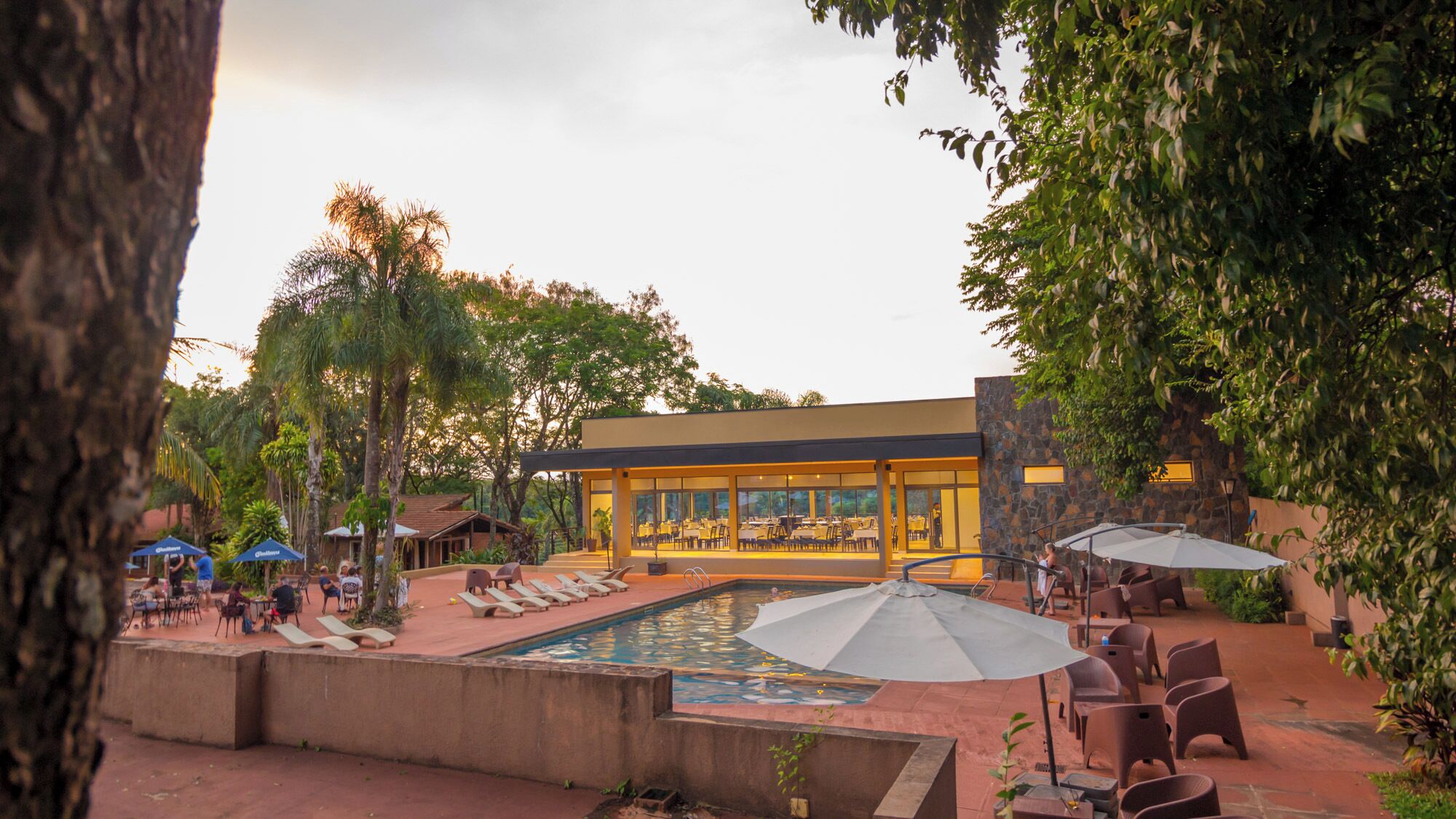 Un'area piscina con lettini e ombrelloni, un ristorante con tavoli all'aperto e un edificio con ampie finestre.