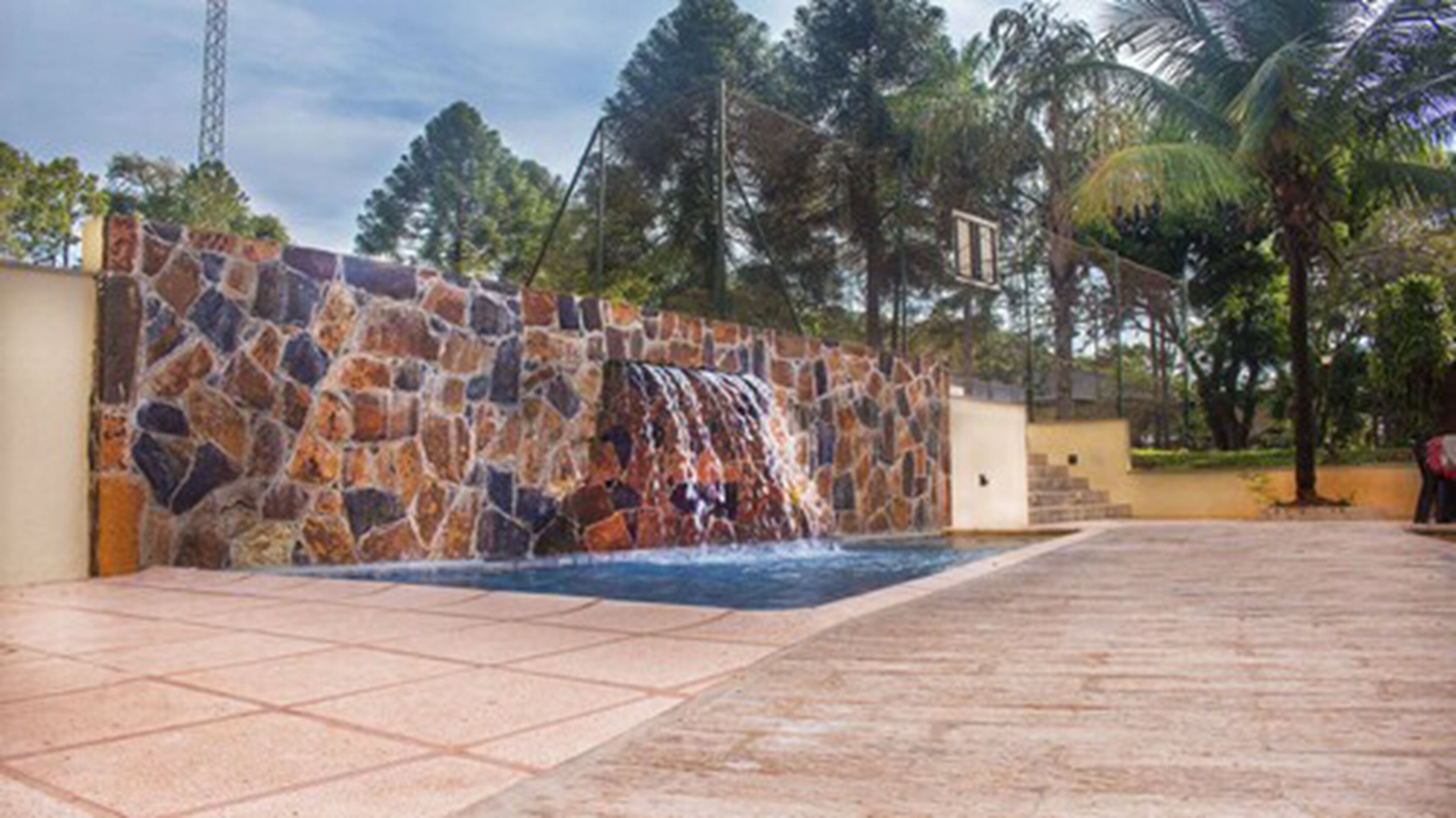 A stone wall with a waterfall feature, a tiled patio, and palm trees in the background.
