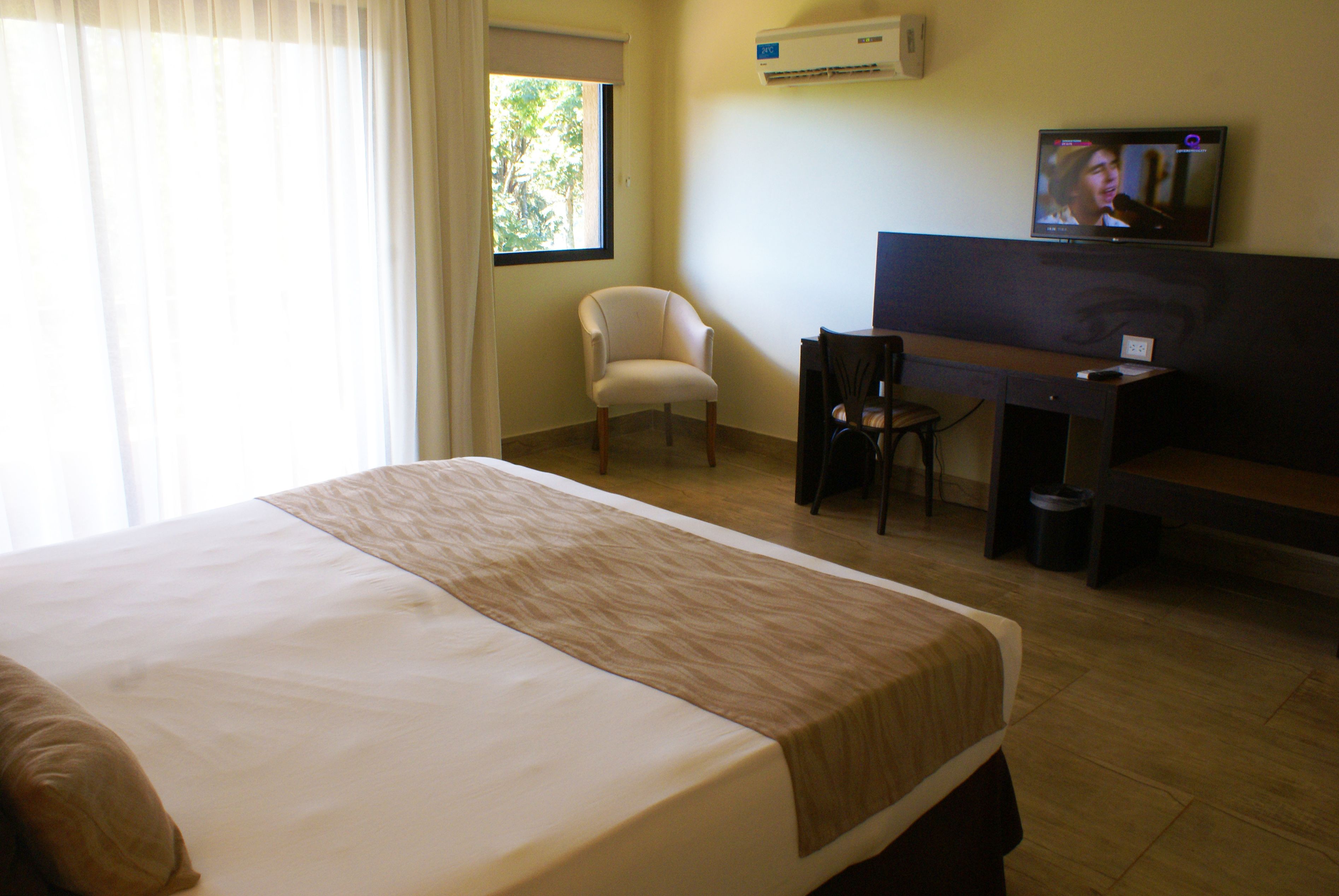 A hotel room with a bed, a desk, a chair, a television, and an air conditioning unit.