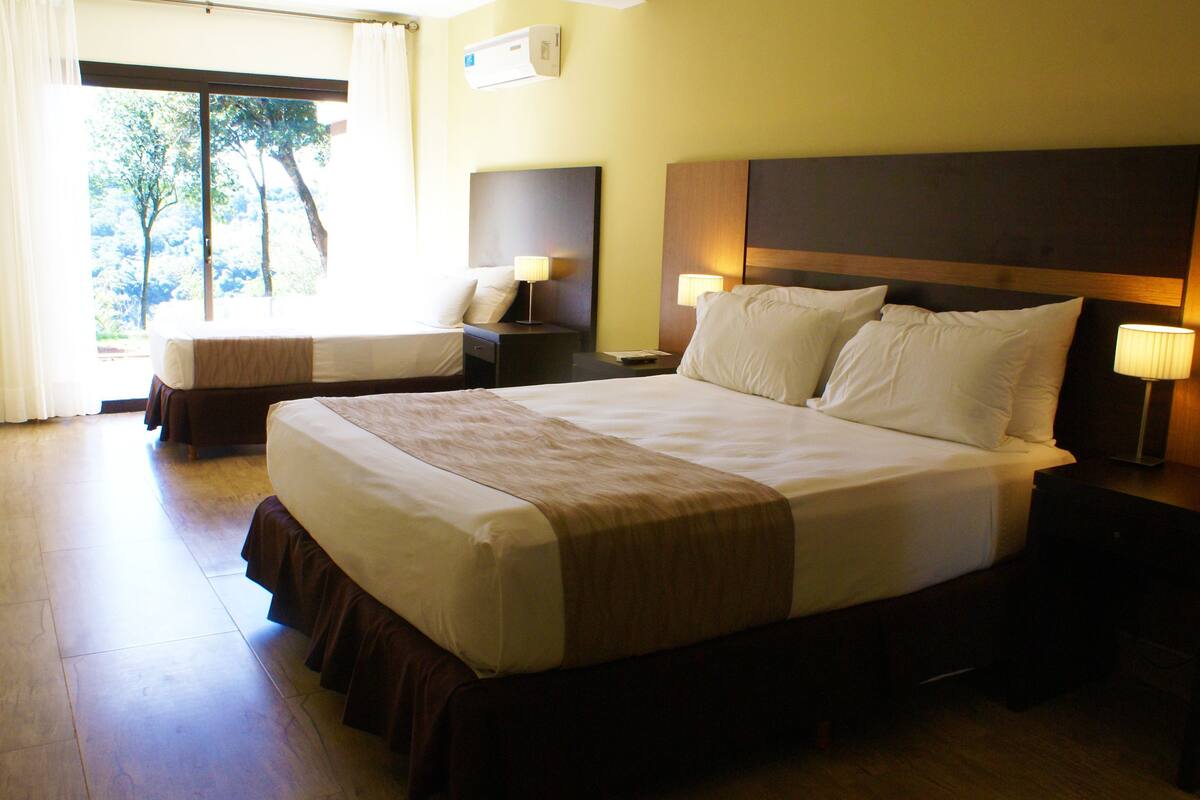 A hotel room with a large bed, two bedside tables with lamps, a window with curtains, and a wall-mounted air conditioner.
