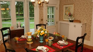 A dining room with a wooden table set for four, surrounded by chairs. There is a chandelier above the table, and a window with a view of greenery outside.