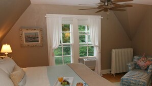 A bedroom with a bed, a window with curtains, a ceiling fan, a radiator, and a chair with a cushion.
