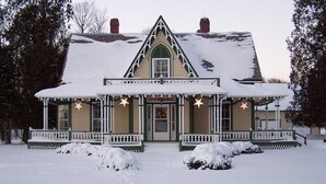 A Victorian-style house with a steeply pitched roof, a front porch with white railings, and a snow-covered front yard.