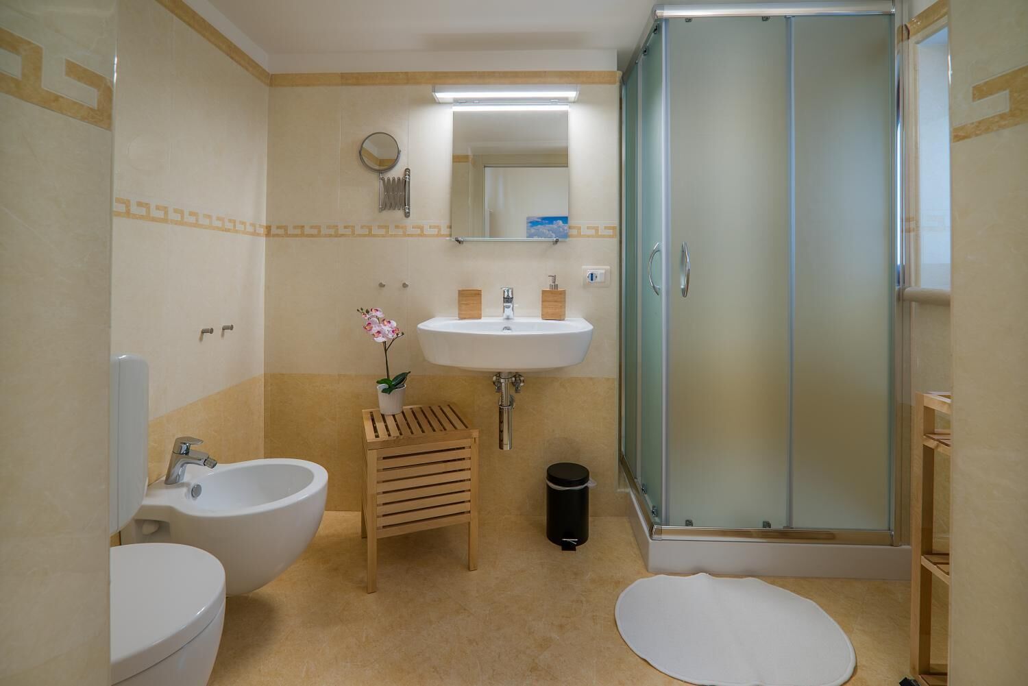 Une salle de bain avec un lavabo, deux toilettes, un petit tabouret en bois, une poubelle et une douche fermée par une paroi de verre.