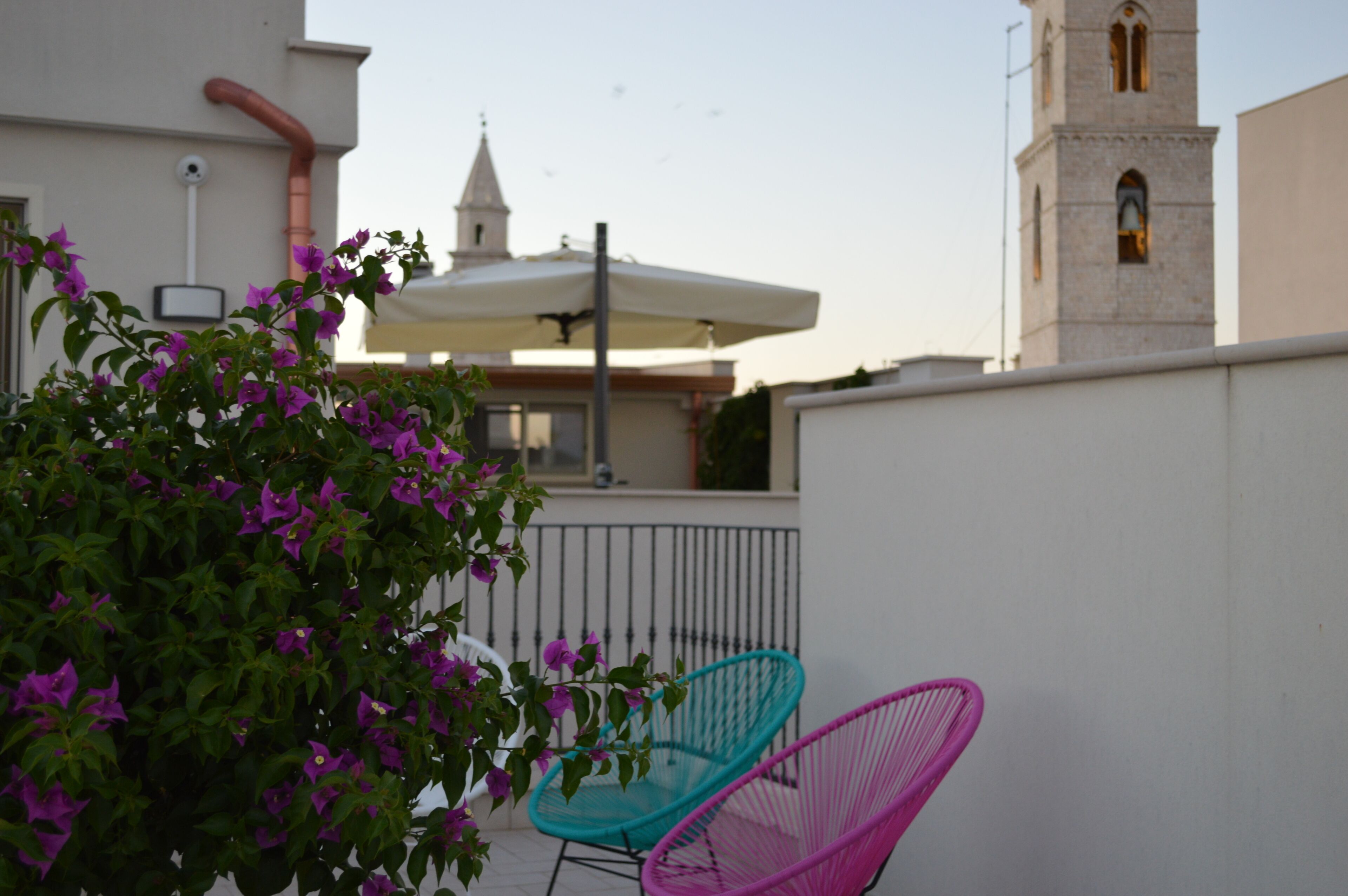 A balcony with a white railing, a turquoise and a pink chair, and a building with a bell tower in the background.
