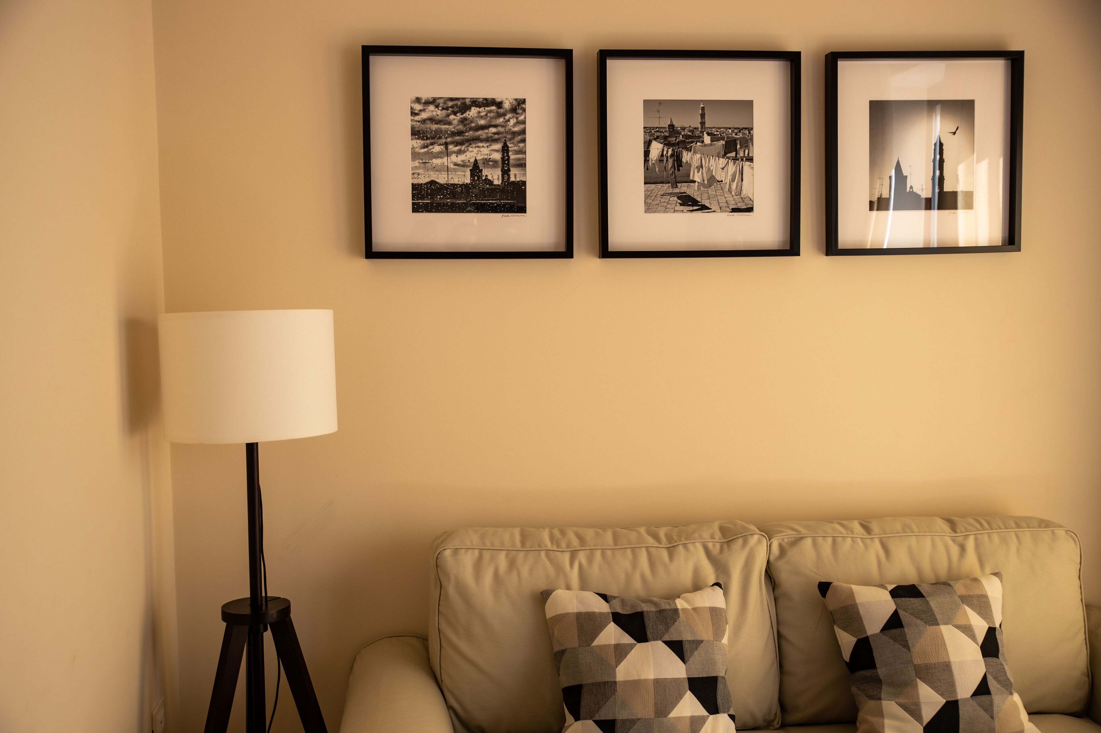 A living room with a beige sofa, two patterned throw pillows, a floor lamp, and three framed pictures on the wall.