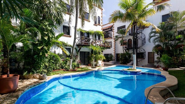 Una piscina rodeada de abundante vegetación y un edificio blanco con balcones.