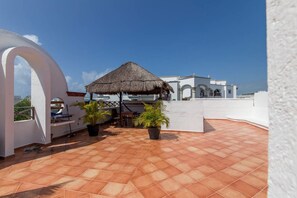 A rooftop terrace with a thatched umbrella, potted plants, and a white building with arches in the background. - Hacienda San Jose by CocoBR (Playa del Carmen)