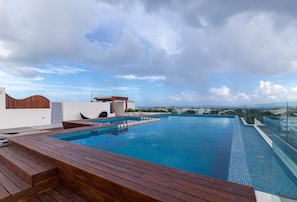 A rooftop infinity pool with wooden deck seating, a glass balustrade, and a view of the cityscape.