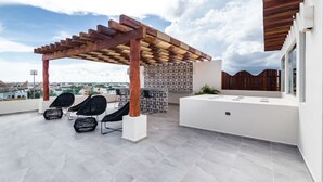 Una terraza en la azotea con una pérgola de madera, sillas de mimbre negro, una zona de comedor y vistas a la ciudad.