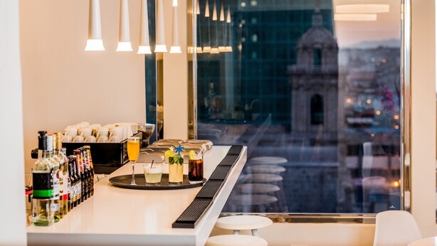 Uma área de bar com garrafas de álcool, um tabuleiro com bebidas e vista para um edifício pela janela.