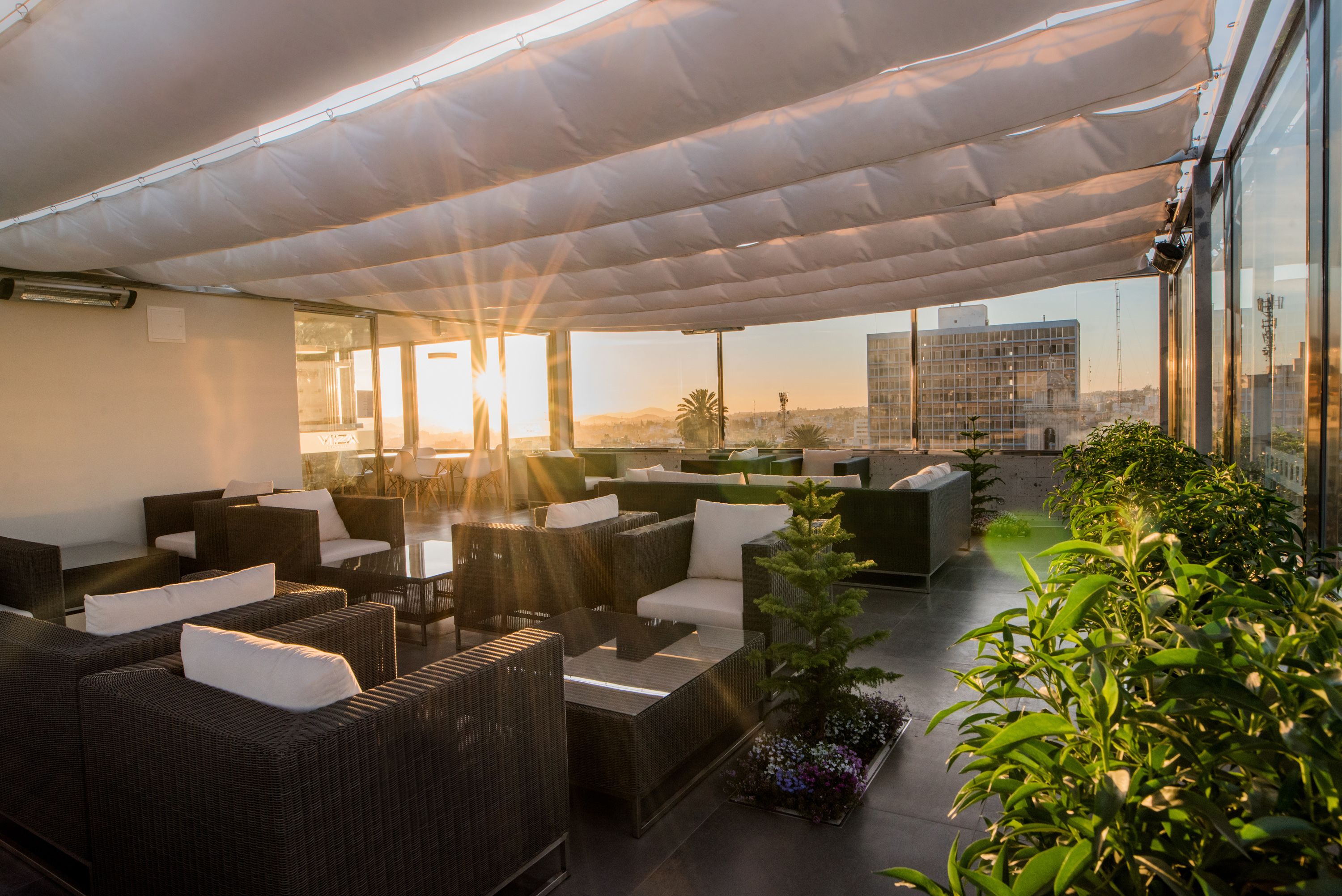 A rooftop lounge with wicker furniture, potted plants, and a view of the cityscape.