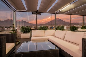 Una terraza en la azotea con sofá, sillas y una mesa, con vistas a un paisaje urbano y montañas.