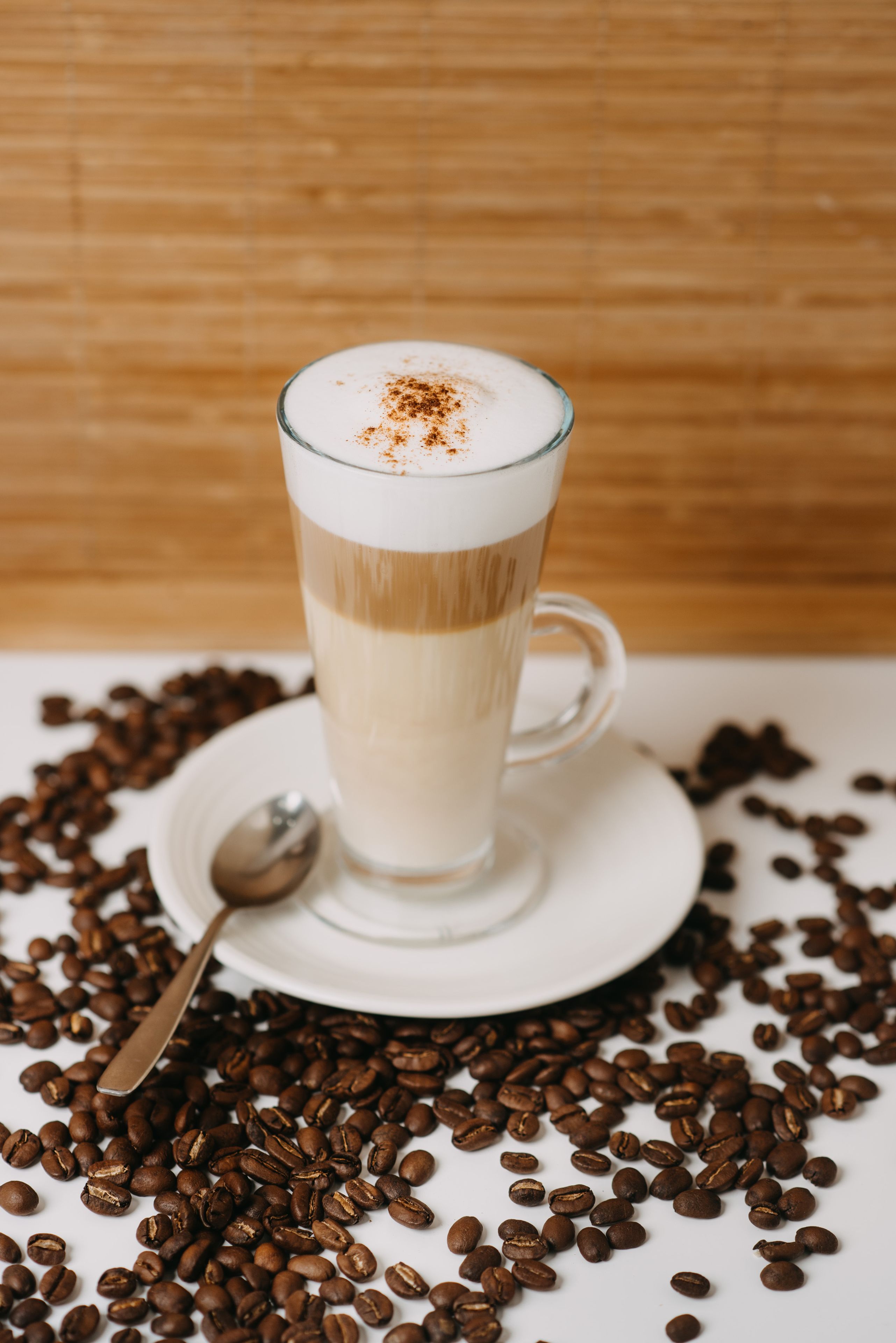 A tall glass of coffee with a frothy top, placed on a saucer surrounded by coffee beans.