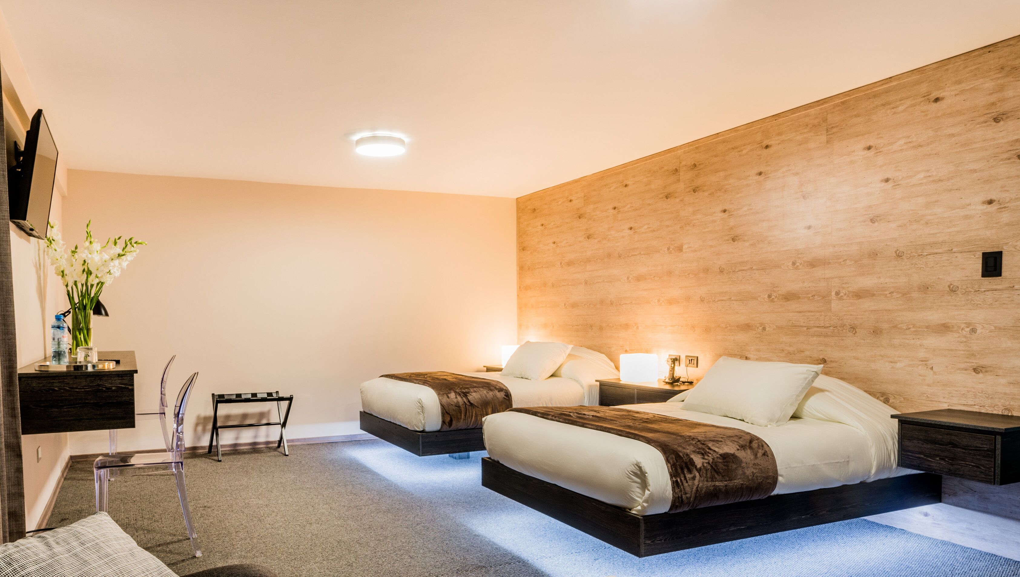 A modern bedroom with two beds, a wooden accent wall, a small desk, and a chair.