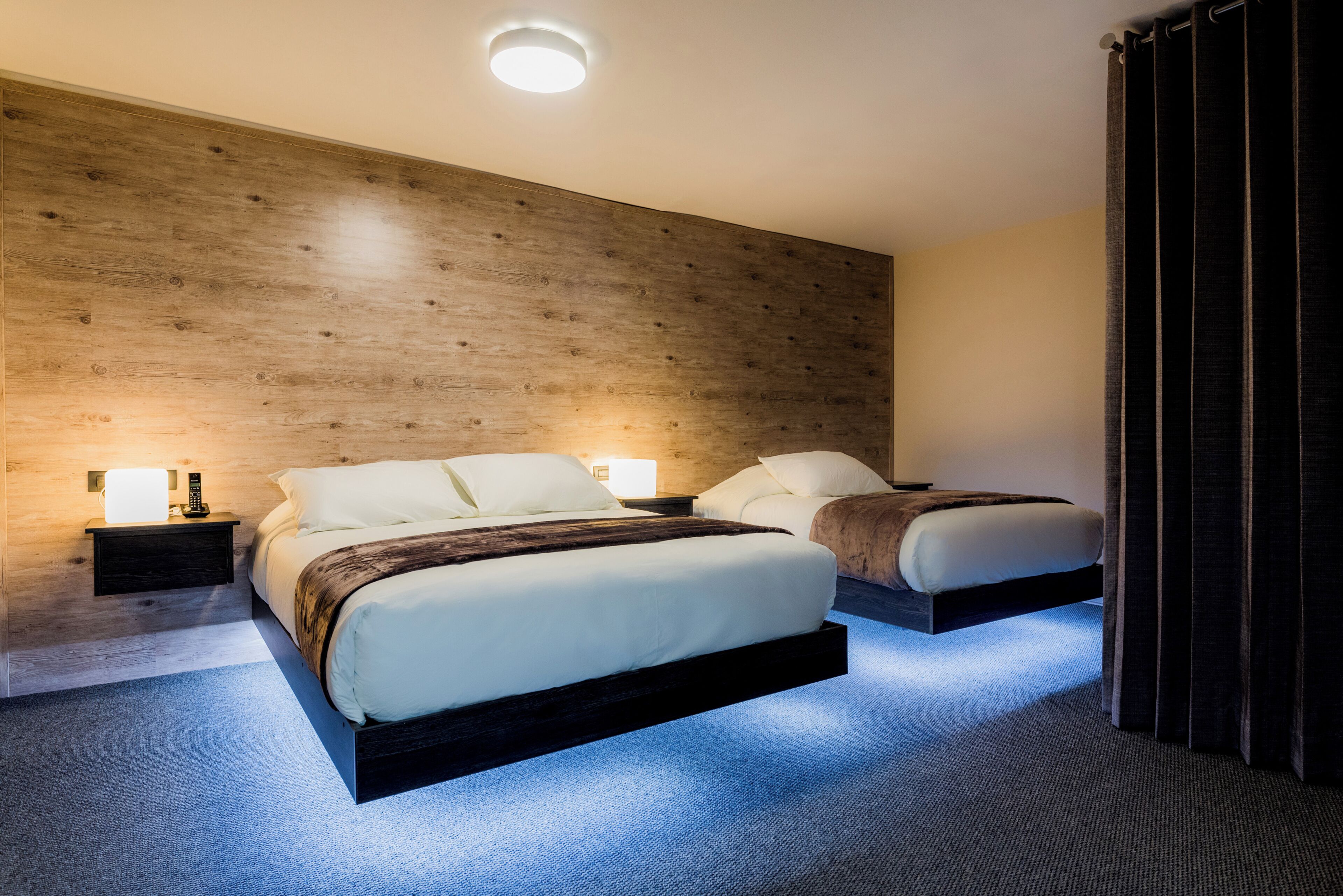 A modern hotel room with two beds, a wooden accent wall, and a blue carpet.