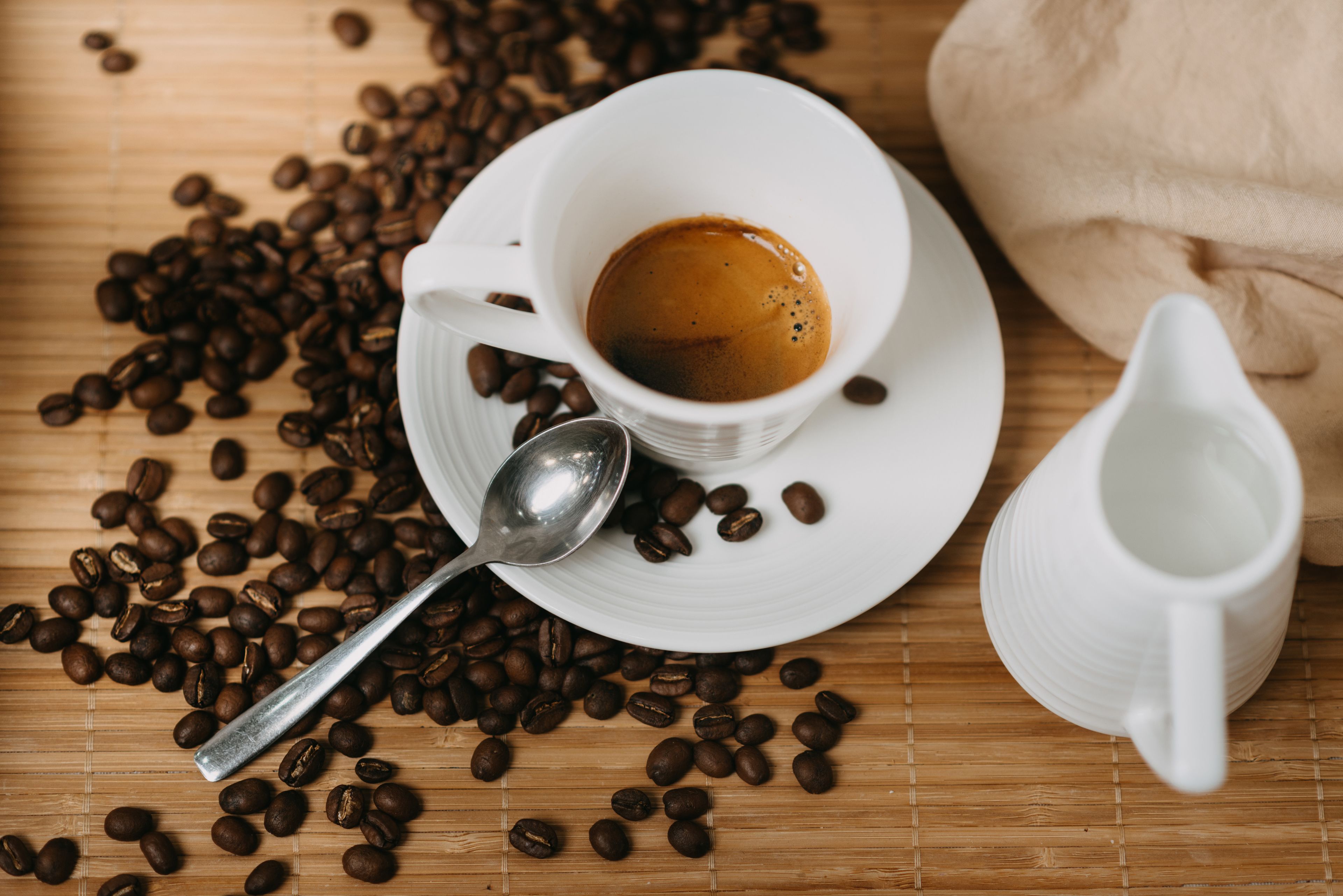 Una taza blanca de café con una cuchara sobre un tapete de bambú, rodeada de granos de café.