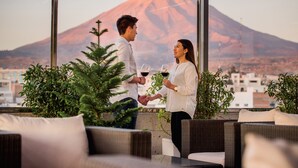 Um casal de mãos dadas brinda com taças de vinho, apreciando a vista das montanhas a partir de uma varanda com móveis de vime e plantas em vasos.