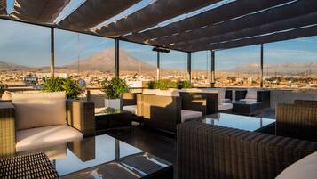 Terraza en la azotea con mobiliario de mimbre, ventanas amplias y vistas a la ciudad y montañas.
