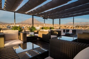 Terraza en la azotea con mobiliario de mimbre, ventanas amplias y vistas a la ciudad y montañas.