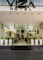 A modern hotel entrance with a glass facade, a visible staircase, and potted plants.