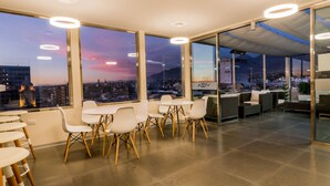 Uma sala de jantar moderna com mesas e cadeiras, grandes janelas com vista para a cidade e um bar com assentos.