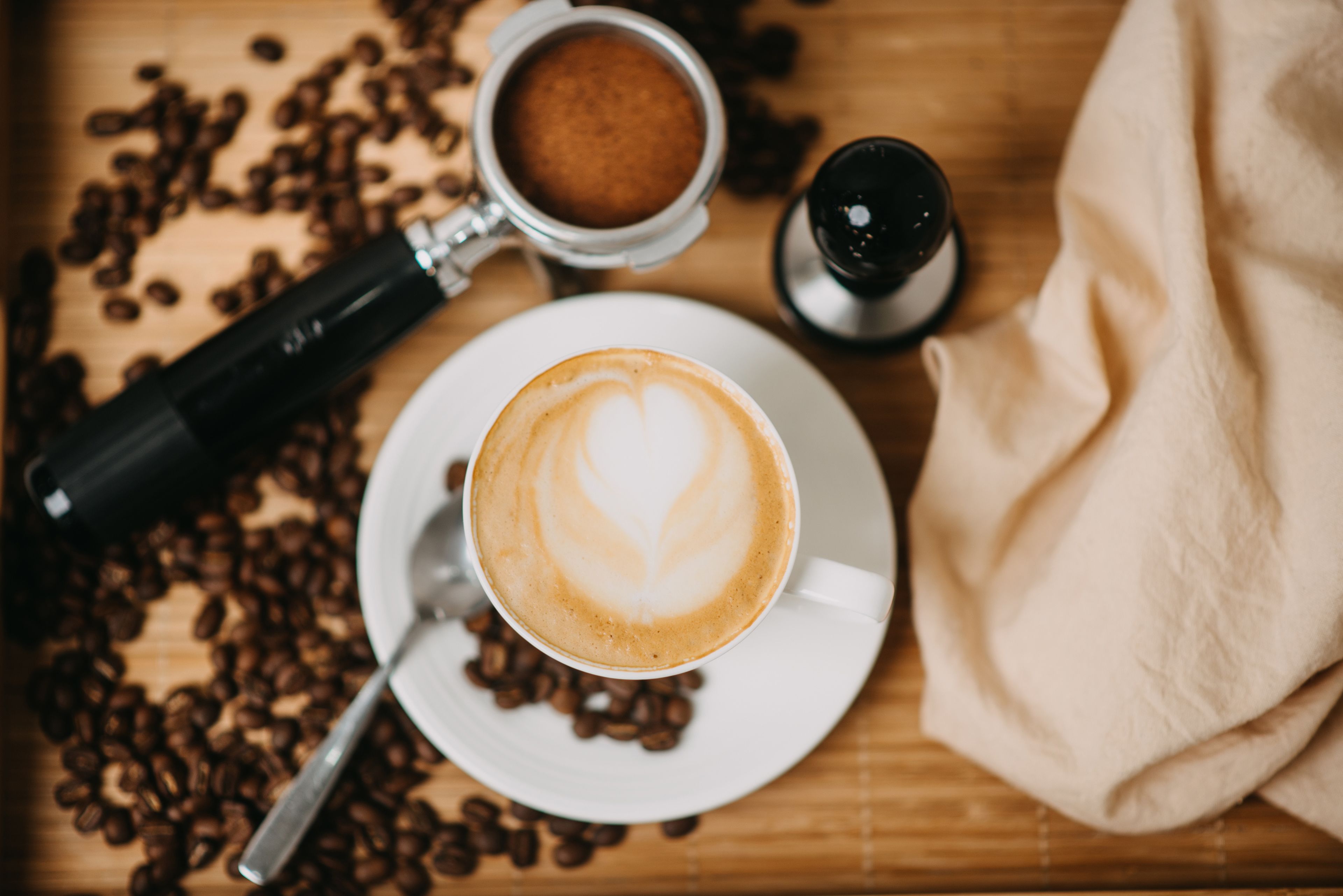 Una taza de café con arte latte sobre un plato, rodeada de granos de café, un molinillo y un salero sobre una superficie de madera.