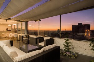 Un salon sur le toit offrant une vue sur la ville au coucher du soleil, avec des meubles en osier et une cloison en verre.