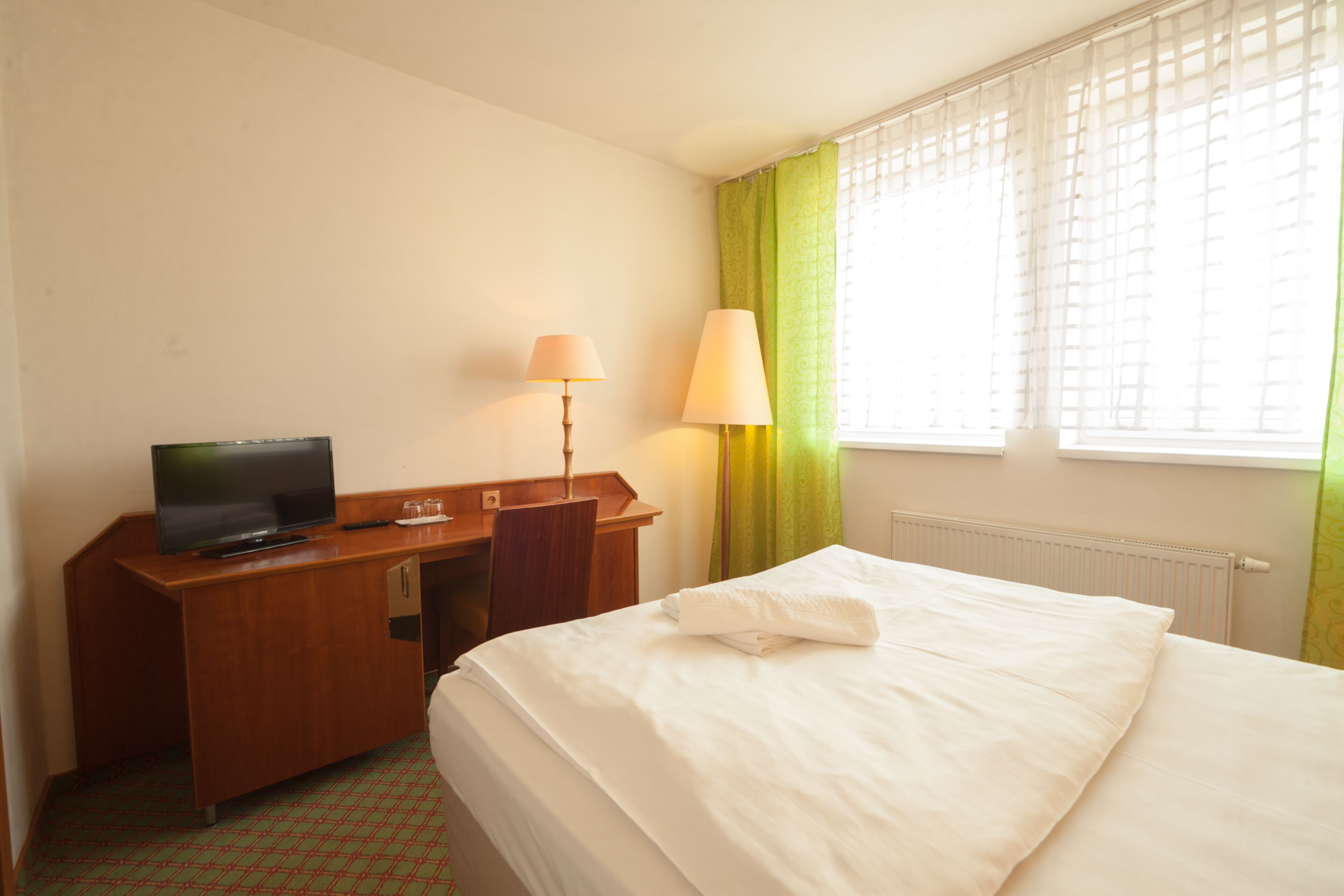 A hotel room with a bed, a desk with a television, a lamp, and a window with curtains.